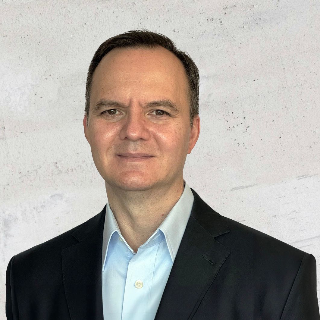 Headshot of Carlos Salazar, Chief AI Officer of Kalosys, wearing a black blazer and white blue shirt smiling lightly against a gray textured background.