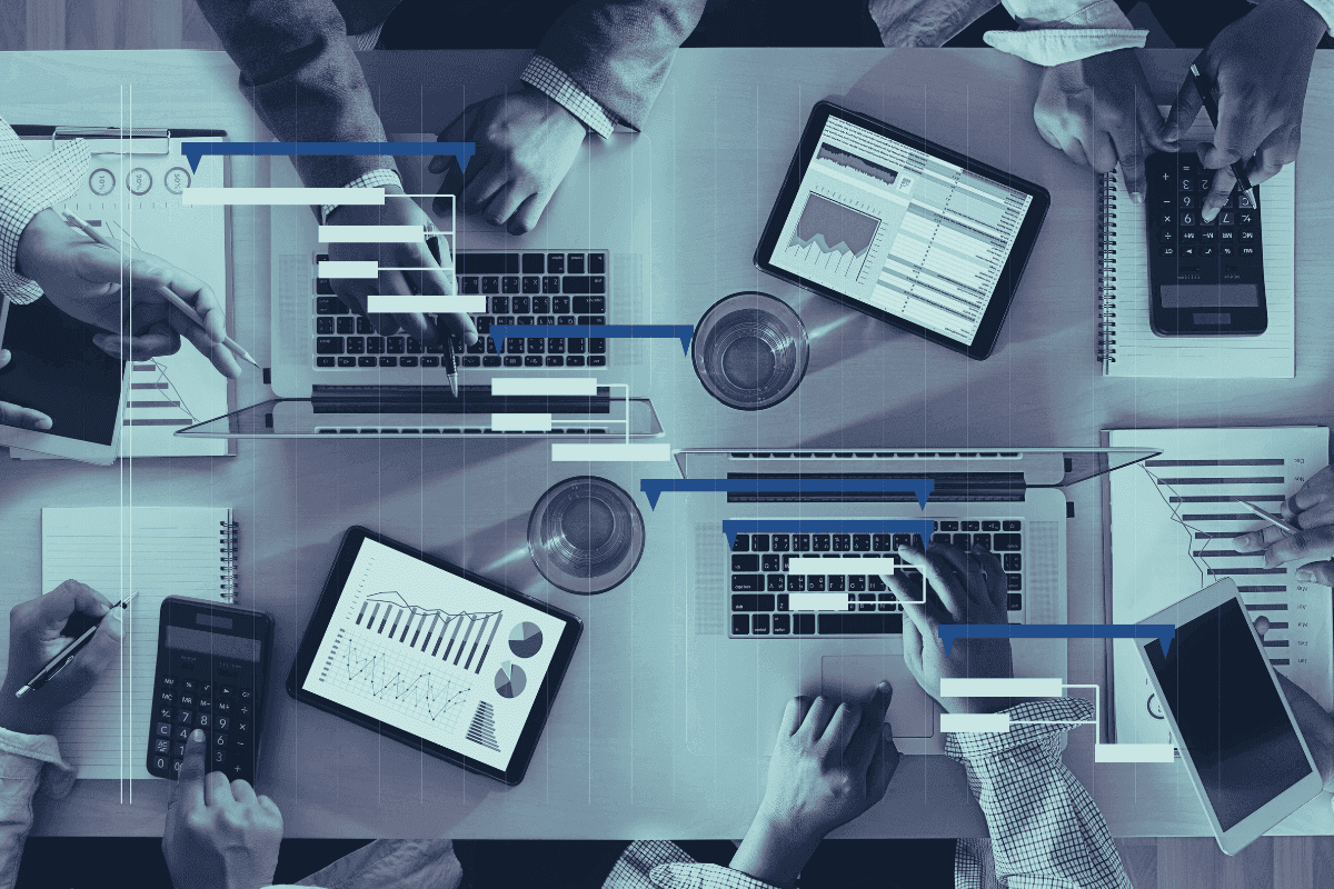 A desk shows multiple tablets and laptops with visual overlays representing the connection of different systems and reports. Multiple hands work on laptops, calculators, and paper across the desk.