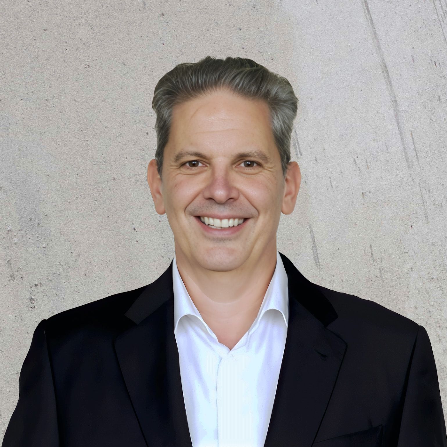 Headshot of Jon Bohlke, Chief Technology Officer, wearing a black blazer and white shirt, smiling brightly against a white concrete background.